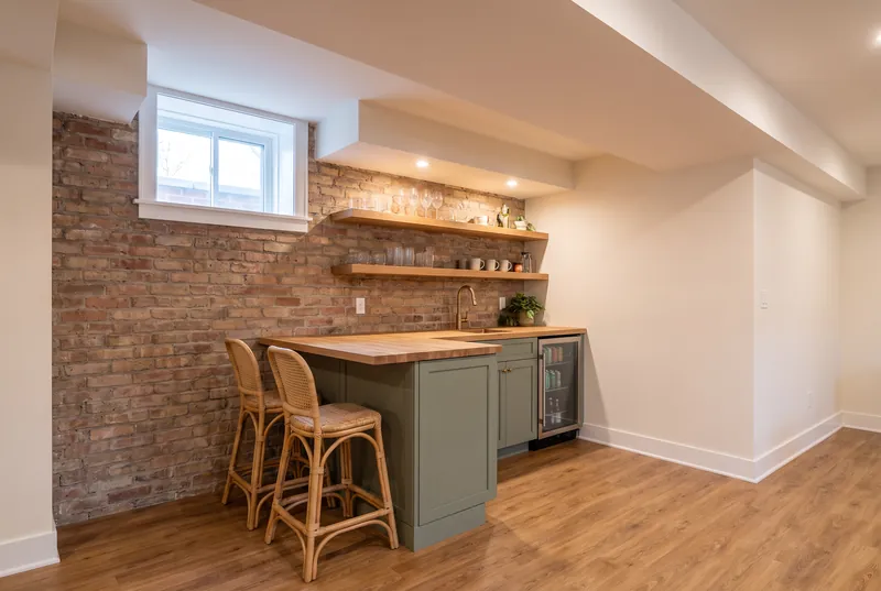 Basement wet bar with sage green cabinets, a butcher block counter and oak shelves on an exposed brick wall