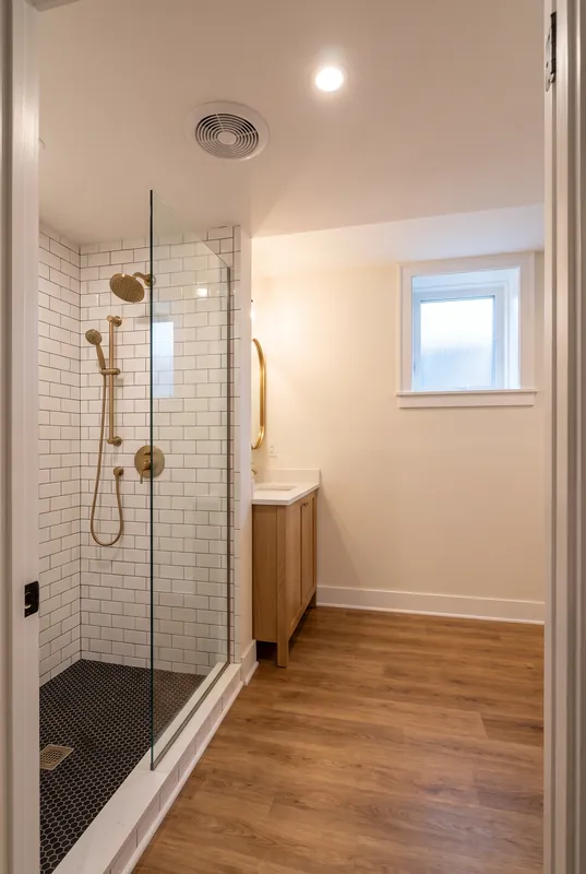 Basement bathroom with a white subway tile shower, a glass panel, black hexagon shower floor tile and brass fixtures