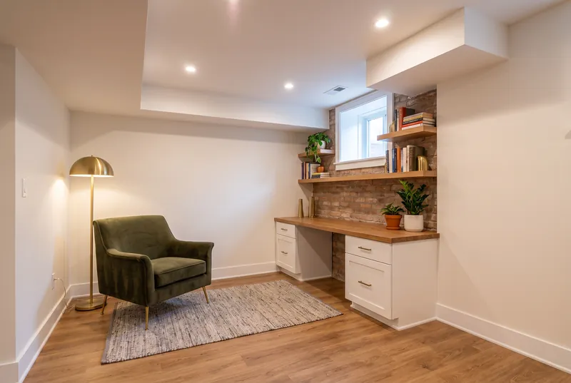 Basement reading and desk nook with a built in desk under a small window, velvet chair, brass floor lamp and oak shelves