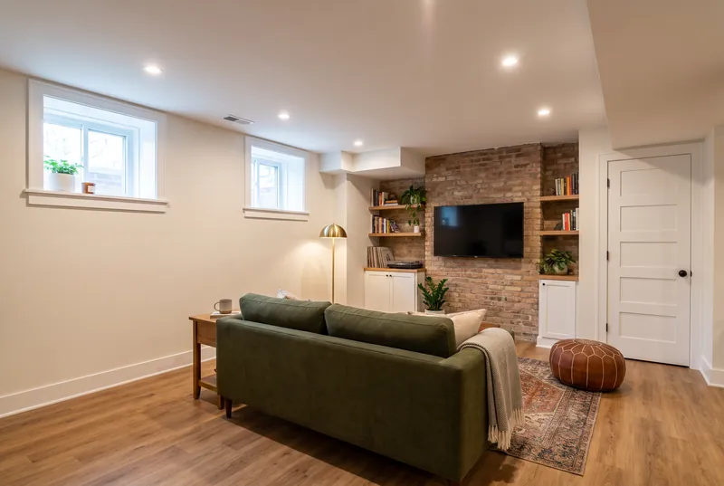 Finished basement family room with an exposed brick wall, olive green sofa, white built in cabinets and oak shelves