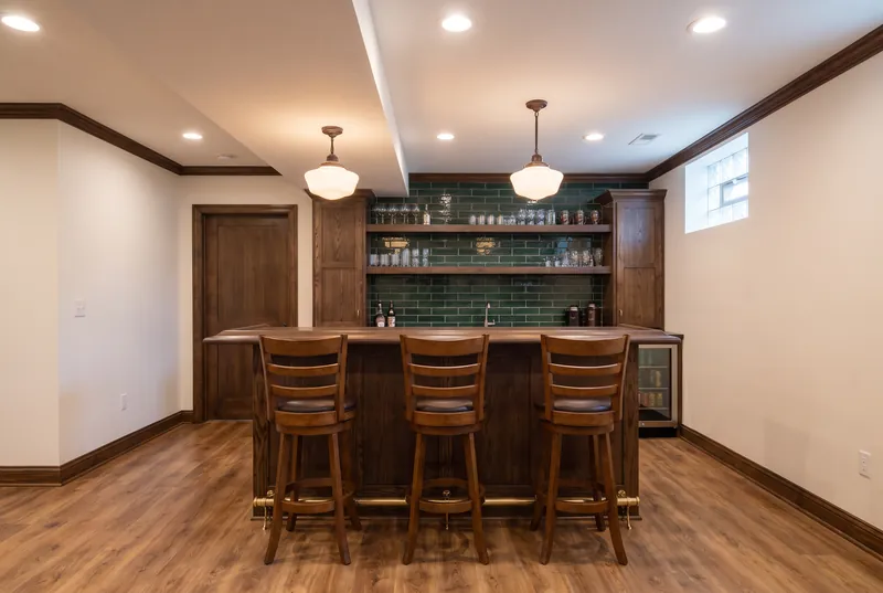 Basement pub bar with a dark wood bar top, brass foot rail, green subway tile and schoolhouse pendants