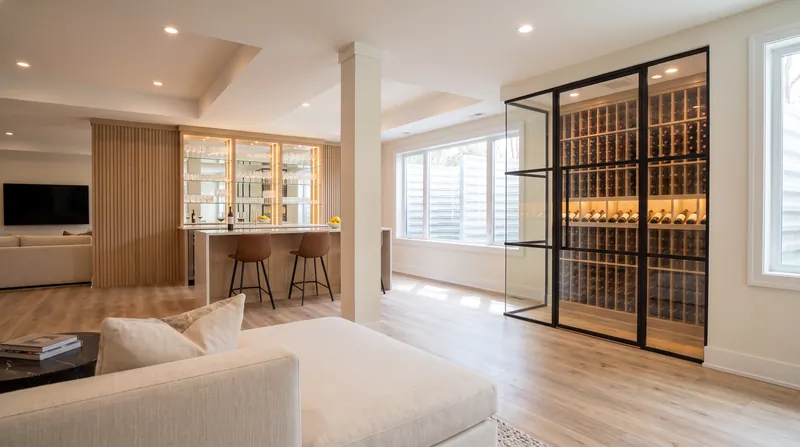 Finished basement lounge with a cream sectional, a white oak bar with leather stools and a black framed glass wine room