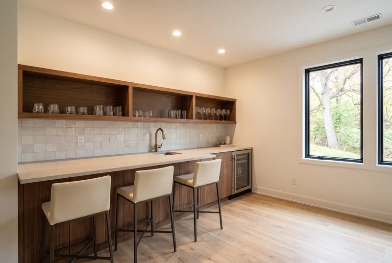 Lower level wet bar with walnut cabinets and open shelves, a stone counter, white tile backsplash and cream bar stools