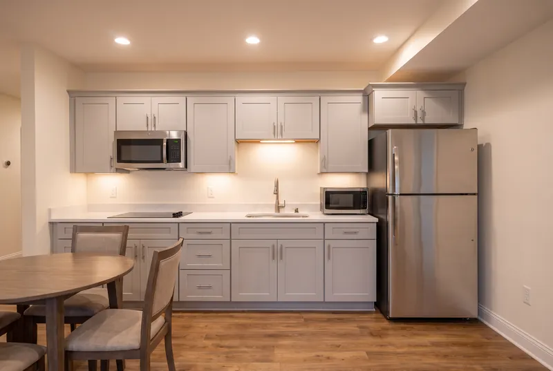 Basement kitchenette with light grey shaker cabinets, a white quartz counter, a cooktop, a microwave and a stainless fridge