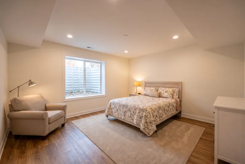 Basement bedroom with an egress window and window well, a bed with a floral quilt and a reading chair