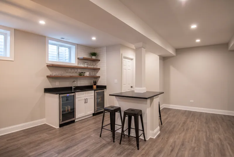 Basement wet bar with white shaker cabinets, a black quartz counter, walnut floating shelves and black metal stools